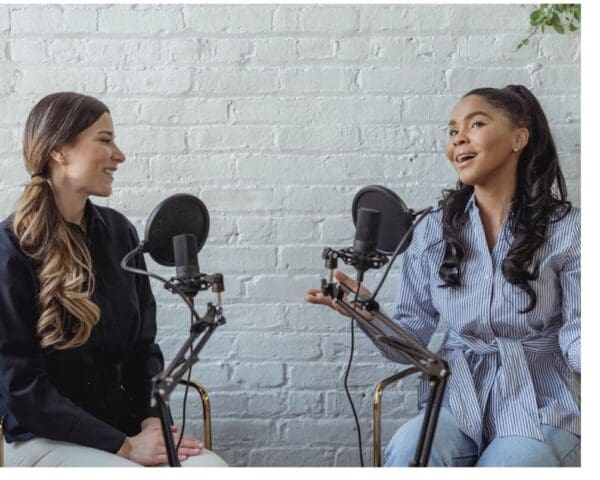 Two women hosting a live podcast, sitting with microphones in front of a white brick wall.