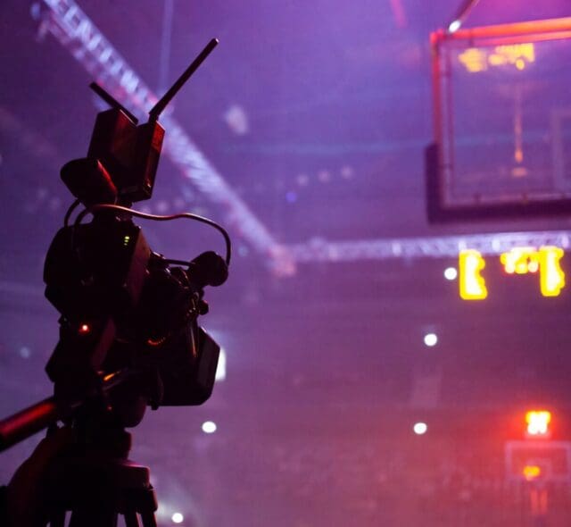 Broadcast camera filming a basketball game with stadium lights and scoreboard in the background.