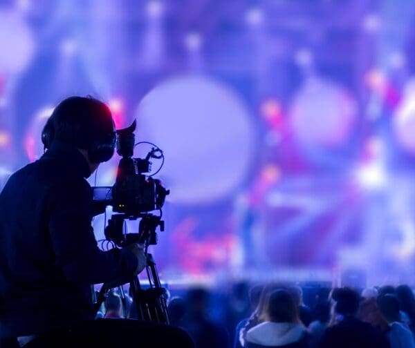 A person filming with a broadcast camera at a live event, with bright lighting and a crowd in the background.