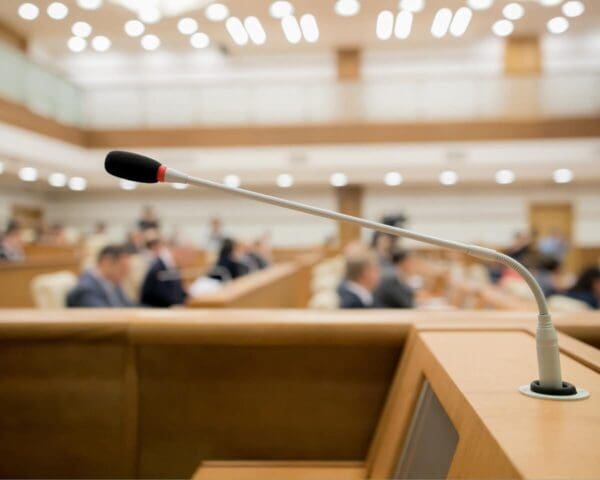 Close-up of a microphone in a government conference room with blurred attendees in the background.
