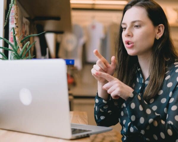 A woman having a video call on a computer in a workspace, speaking and gesturing with her hands.