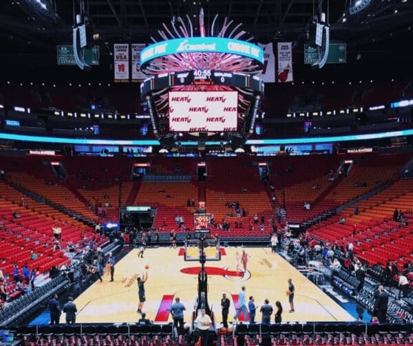 Wide shot of an empty basketball stadium with players warming up and a large scoreboard overhead.